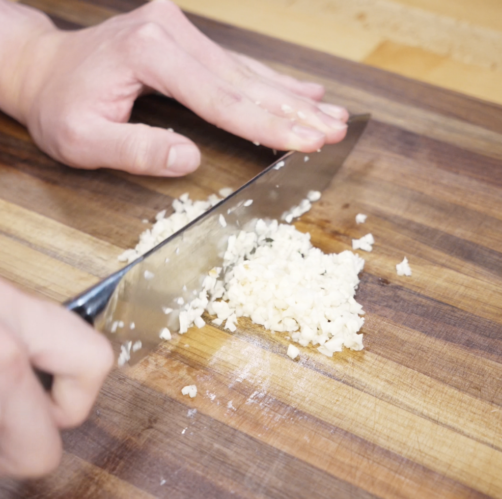 A chef minces a generous amount of garlic on a wooden cutting board, preparing the key aromatic ingredient for the honey garlic sauce.