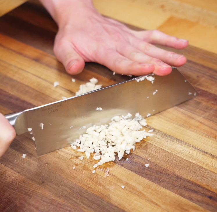 Garlic cloves are finely minced on a wooden cutting board with a sharp knife, ensuring maximum flavor in every bite.