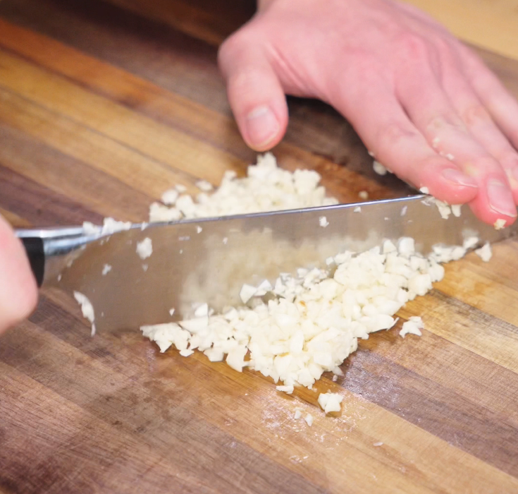 Garlic cloves are finely minced on a wooden cutting board, creating the bold aromatic base for the noodles.