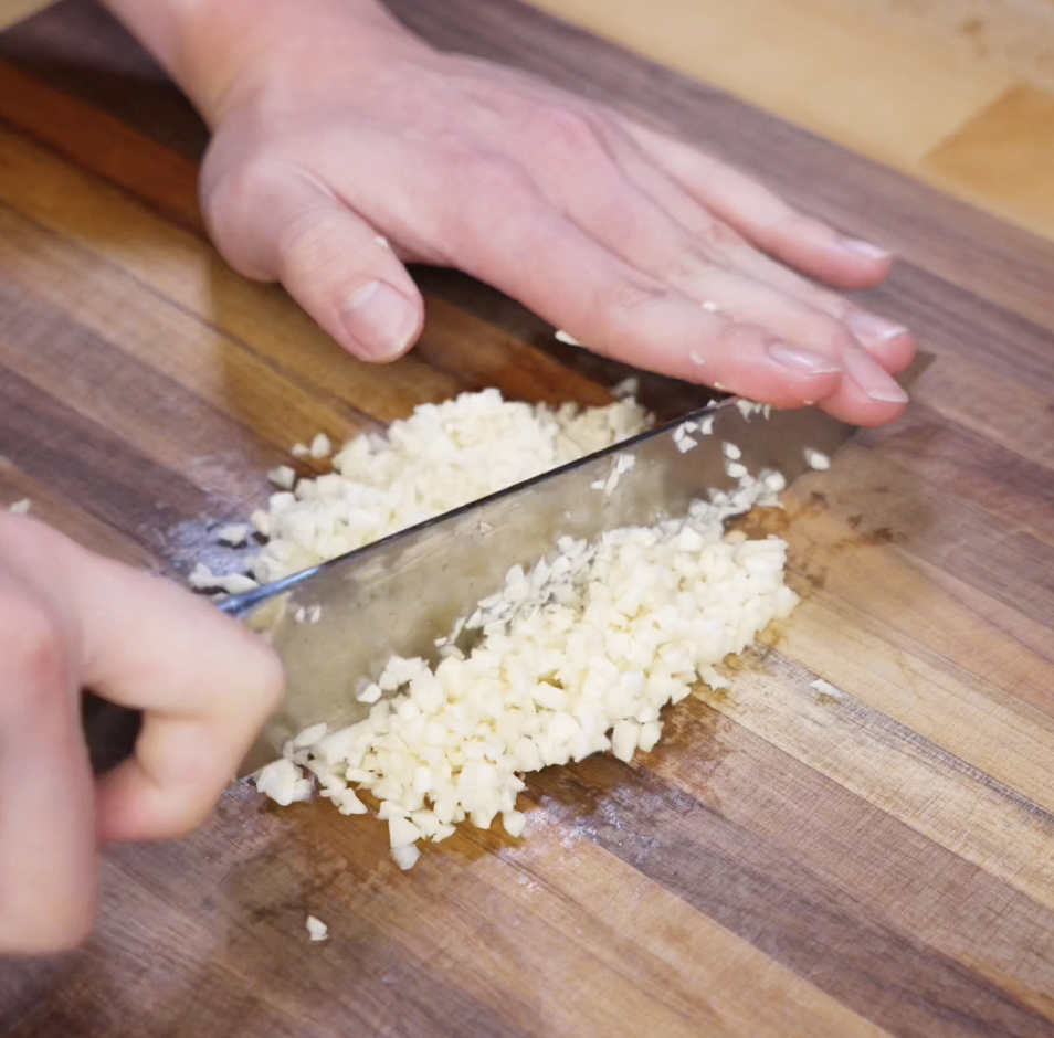 A chef’s knife rapidly chops fresh garlic cloves into a fine mince on a wooden cutting board, preparing for crispy garlic fried rice.