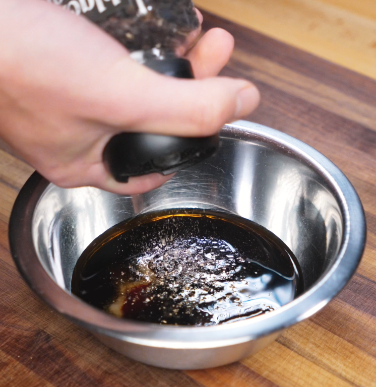 A hand grinds fresh black pepper into a metal bowl filled with soy sauces, oyster sauce, sesame oil, and vinegar — the savory base of the black pepper chicken sauce.
