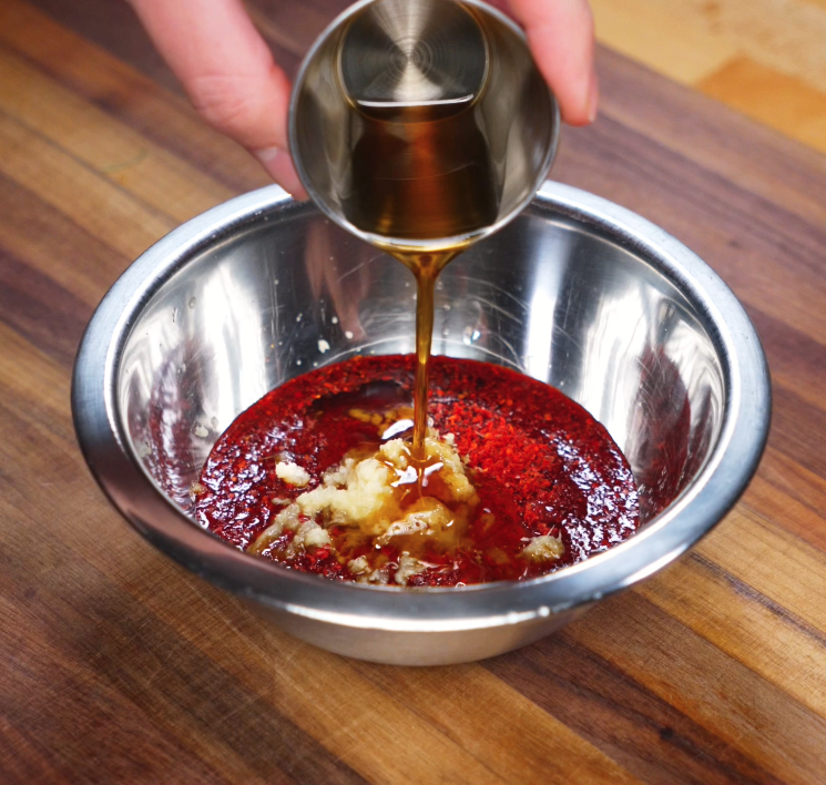 Sesame oil is poured into a bowl of red gochujang, garlic, ginger, and chili flakes to form a spicy, savory-sweet marinade for pork bulgogi.