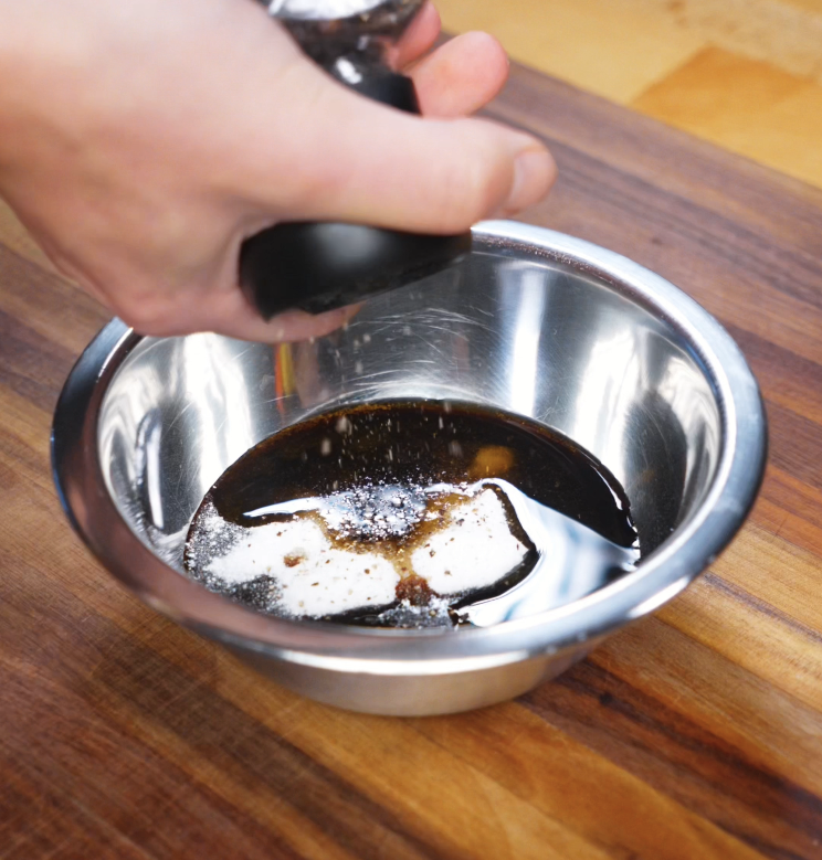 A hand grinds black pepper into a stainless steel bowl filled with soy sauces, sesame oil, oyster sauce, and sugar to create the savory sauce.