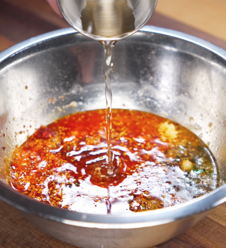 A stream of rice vinegar is poured into a metal bowl of spicy peanut chili oil mixture. This step balances out the richness with tang and umami, completing the noodle sauce.