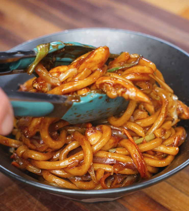 A pair of teal tongs places a serving of glossy chicken yaki udon into a black bowl. The chewy noodles, stir-fried with tender chicken, onions, and carrots, are coated in a rich soy-based sauce.