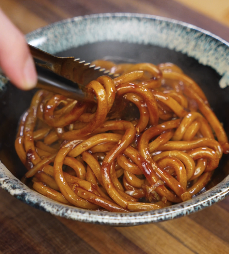 A pair of tongs lifts a generous portion of sticky, saucy udon noodles into a bowl, ready to be served hot.