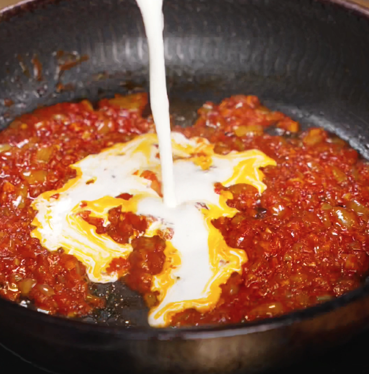 A steady stream of heavy cream swirls into a simmering tomato-gochujang mixture, transforming the sauce into a luscious, spicy cream blend.