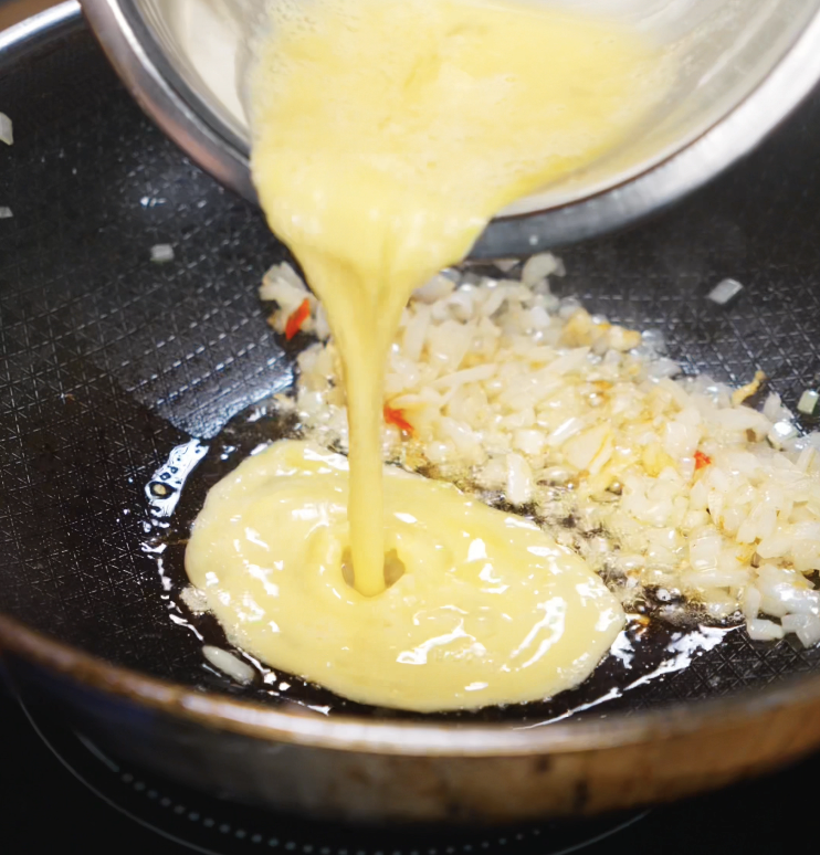 Whisked eggs are poured into the pan alongside garlic, chilies, and onion. The scrambled eggs will add richness and texture to the dish.