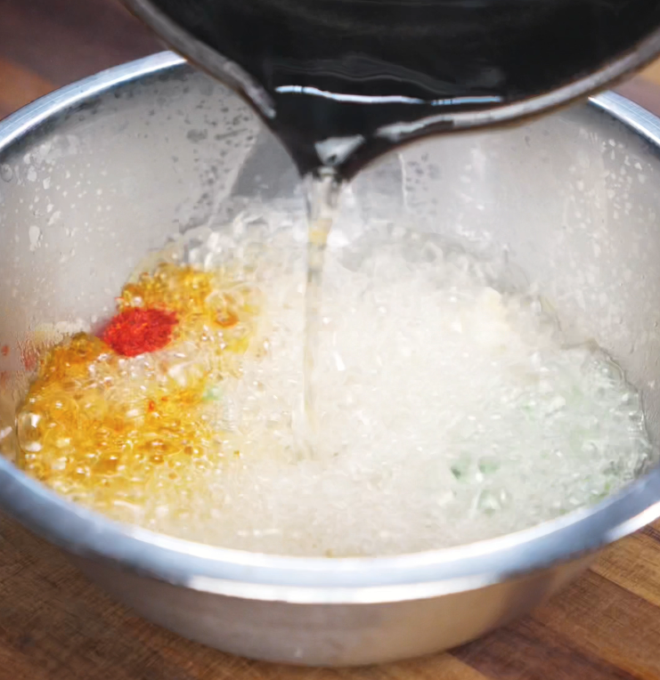 Bubbling hot oil is poured into a metal bowl filled with peanut butter, scallions, garlic, and chili flakes. The sizzling bloom releases deep flavor and aroma for the noodle sauce.