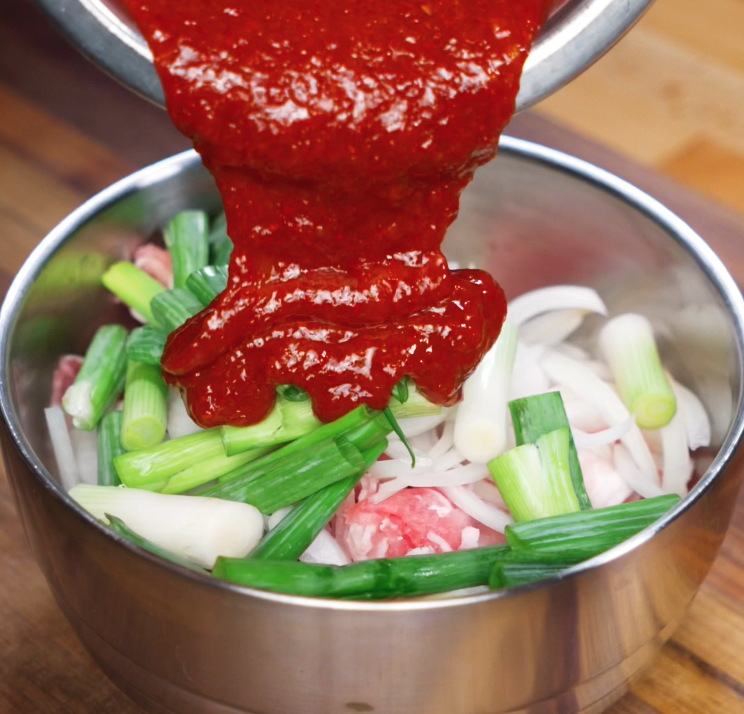 A bold red gochujang marinade is poured over thinly sliced pork, onions, and scallions in a metal mixing bowl, ready for marination.