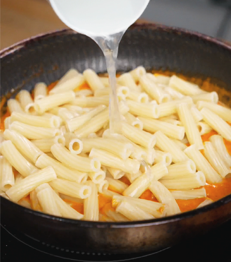 Reserved starchy pasta water is poured into a pan of cooked rigatoni, helping the spicy cream sauce cling perfectly to every noodle.