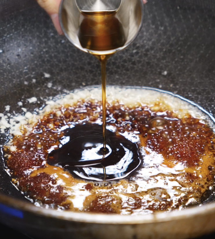 A stream of soy-based sauce is poured into a pan of sautéed garlic and butter to create a sweet, savory glaze for the chicken.