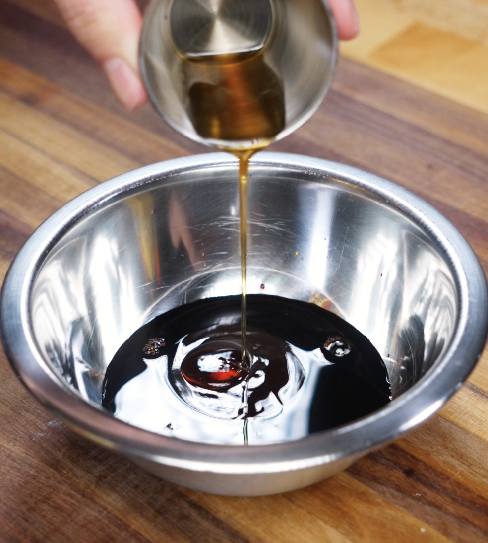 A golden stream of sesame oil is being poured into a metal bowl of soy sauce, oyster sauce, and seasonings to form the glossy stir fry sauce.