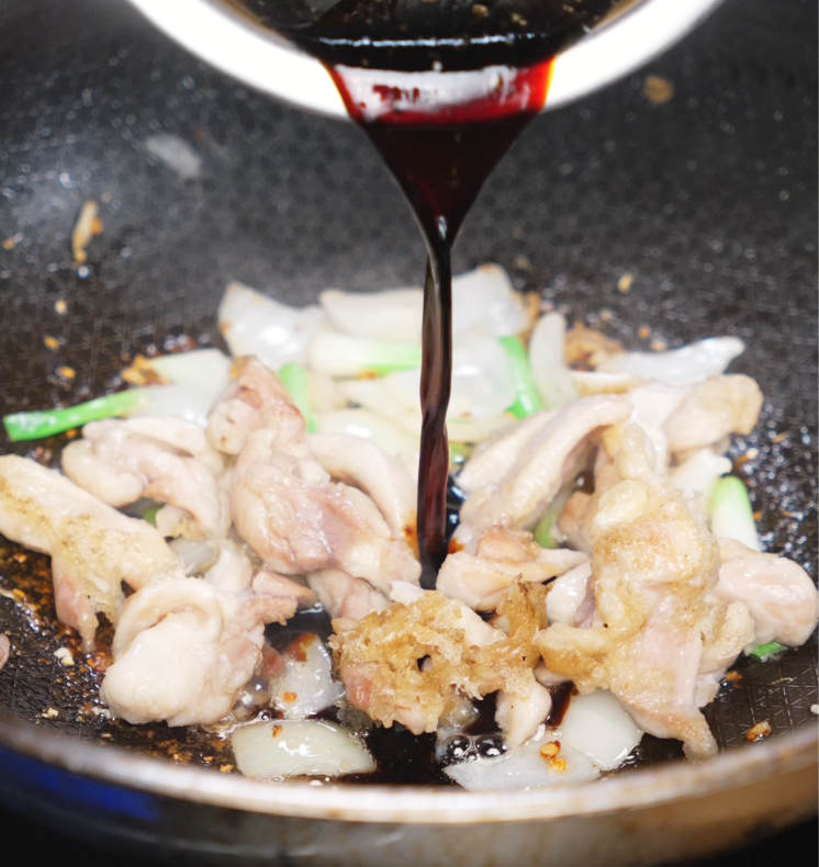 A glossy black pepper sauce is poured over browned chicken, garlic, and onions in a skillet to coat everything in bold flavor.