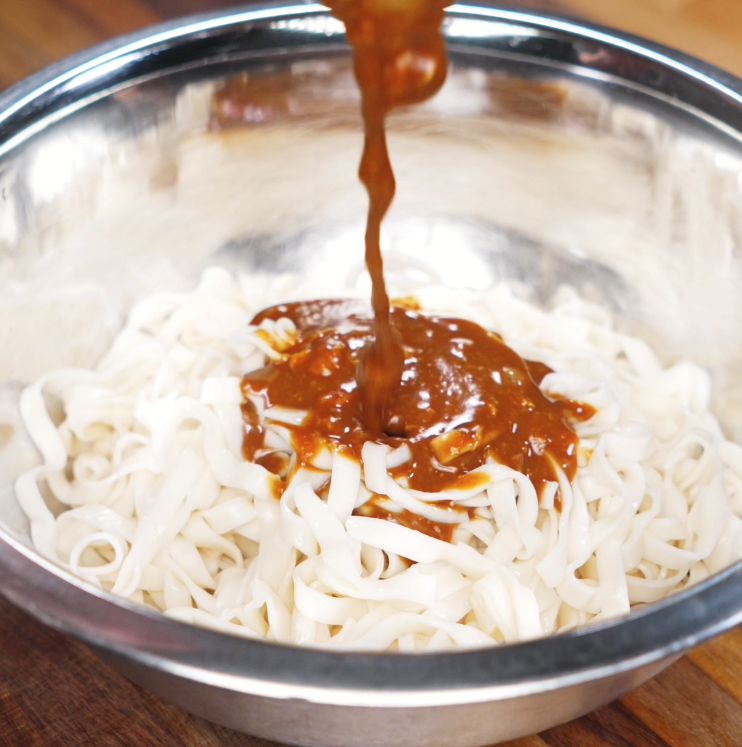 A glossy peanut-sesame sauce is poured over freshly boiled noodles in a mixing bowl, ready to be tossed together.