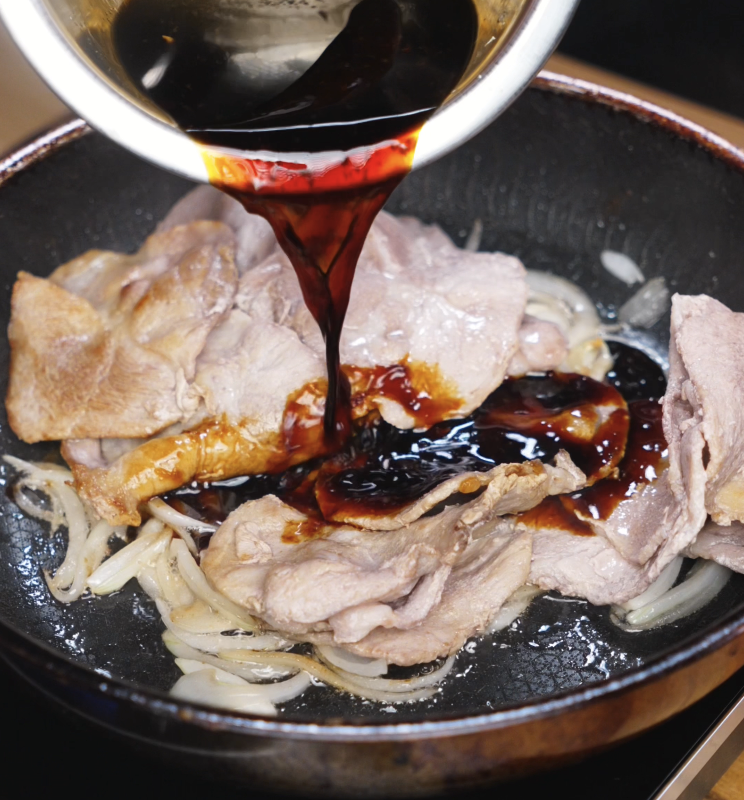 A glossy soy-based sauce is poured over browned pork and sautéed onions in a hot pan, beginning to simmer.