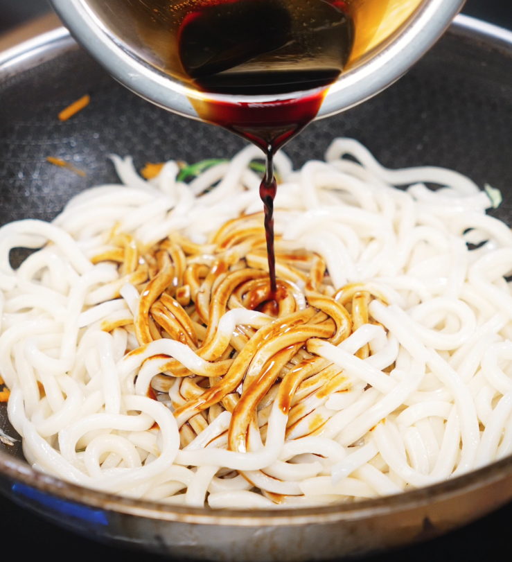 A soy-based sauce is poured over freshly boiled udon noodles in a pan filled with stir-fried veggies, ready to be tossed with chicken.
