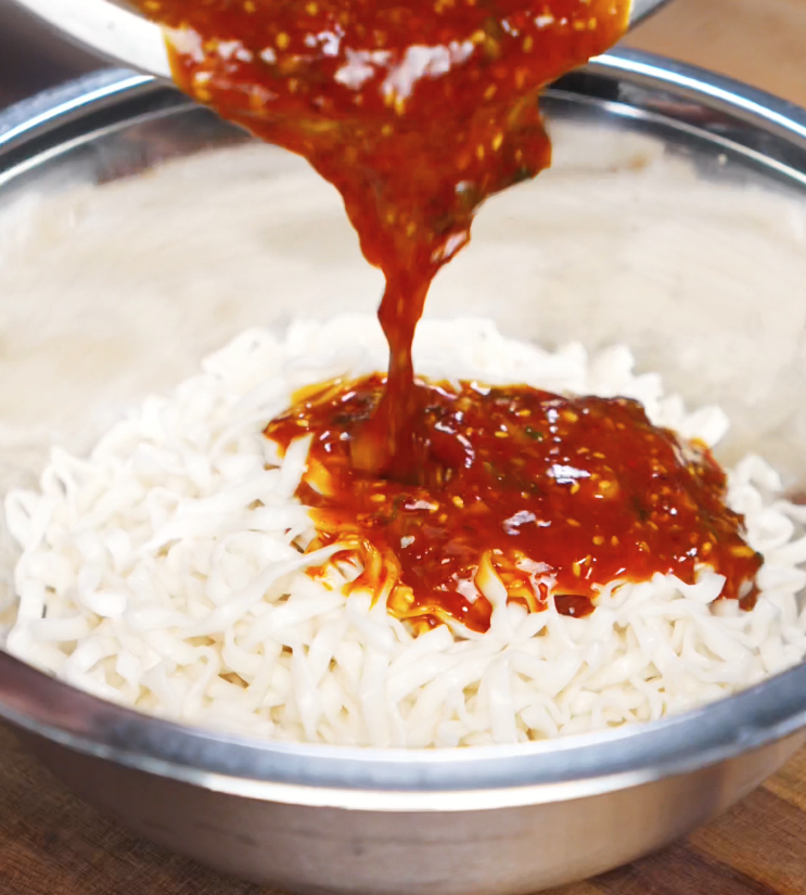 A bright red chili oil peanut sauce is poured over freshly boiled noodles in a metal bowl. The thick, glossy mixture will coat each strand for maximum flavor.