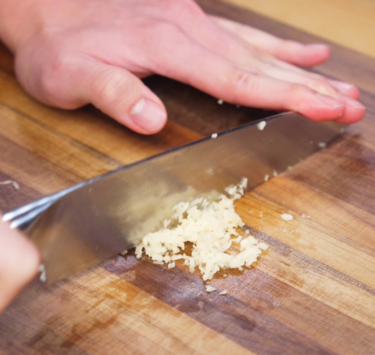 A chef’s knife minces fresh garlic on a wooden cutting board. The finely chopped garlic will be bloomed in hot oil to form the flavorful base of these peanut chili oil noodles.