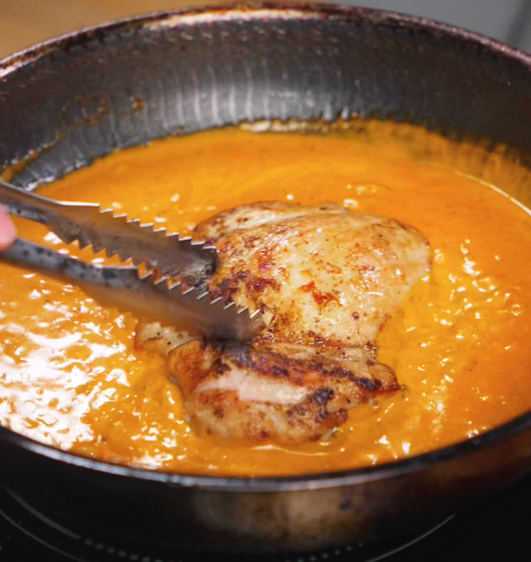 A seared chicken thigh is lowered into the creamy tomato-gochujang sauce, ready to soak up all the spicy richness before slicing.