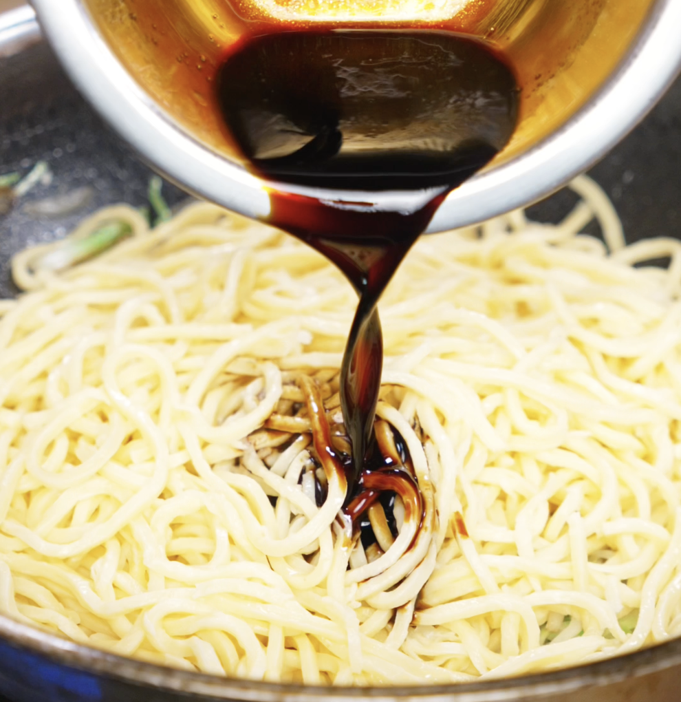 A rich, dark soy-based sauce is being poured over just-cooked noodles in the wok. The sauce will coat the noodles and tie all the flavors together.