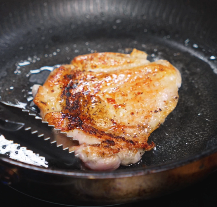 A marinated chicken thigh sizzles skin-side down in a hot skillet, developing a deep, golden brown crust for maximum flavor.
