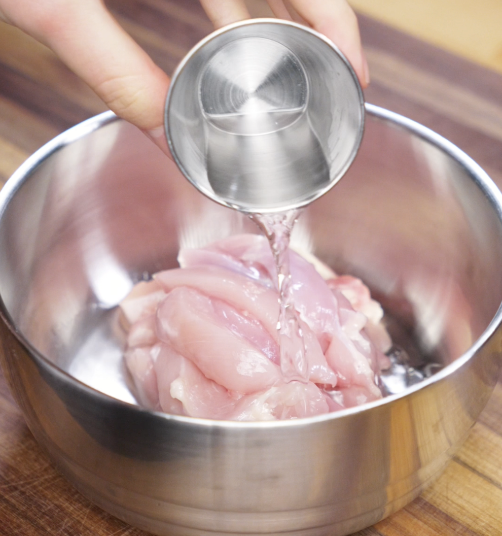 A hand pours Shaoxing wine over raw sliced chicken thighs in a metal bowl. This step helps tenderize the meat and infuse it with classic Chinese flavor.