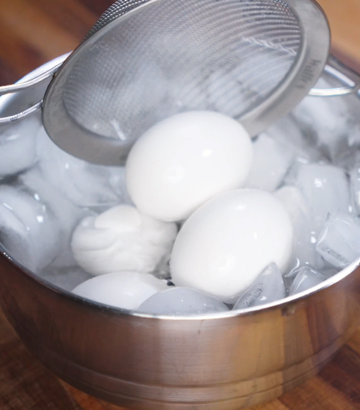 Hard-boiled eggs are transferred into a bowl of ice water with a strainer to halt cooking and make peeling easier.