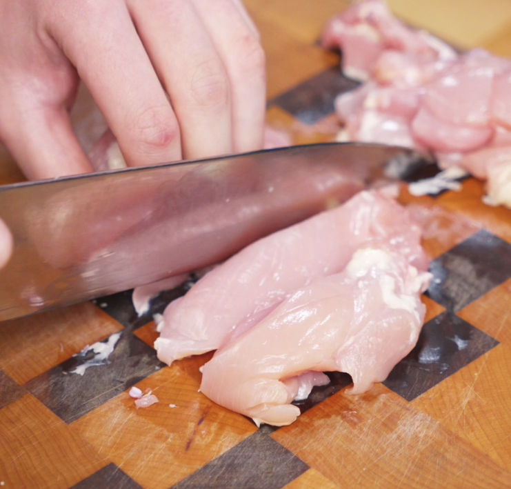 A hand uses a knife to slice boneless chicken thighs into even pieces on a woodgrain cutting board for the stir-fry.