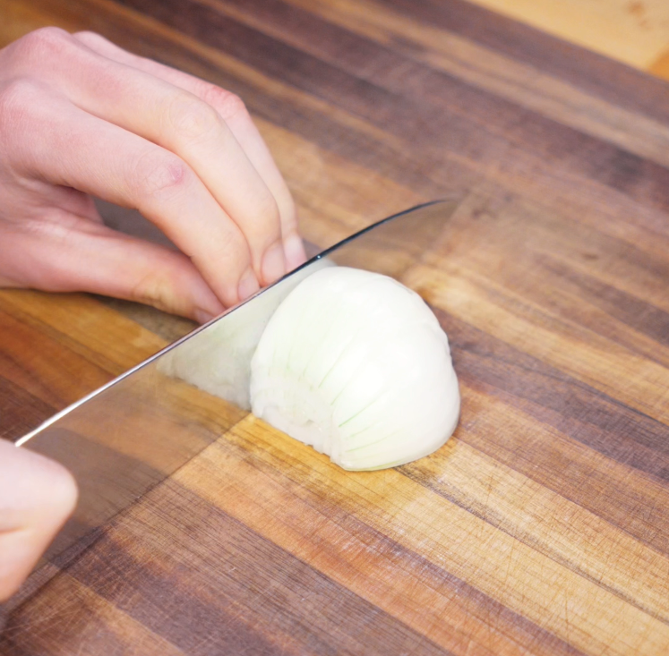 A chef’s knife cuts a white onion into wedges on a wooden cutting board to prep for the stir-fry base.