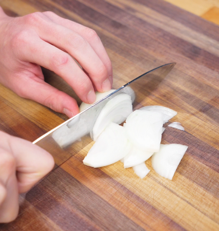 A hand uses a chef’s knife to thinly slice half an onion on a wooden cutting board for the vegetable base of the stir-fry.