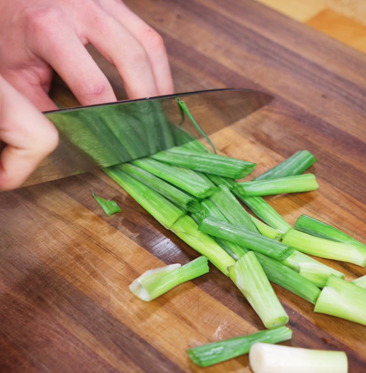 A hand slices fresh scallions into ½-inch pieces on a wooden cutting board, prepping aromatics for the bulgogi marinade.