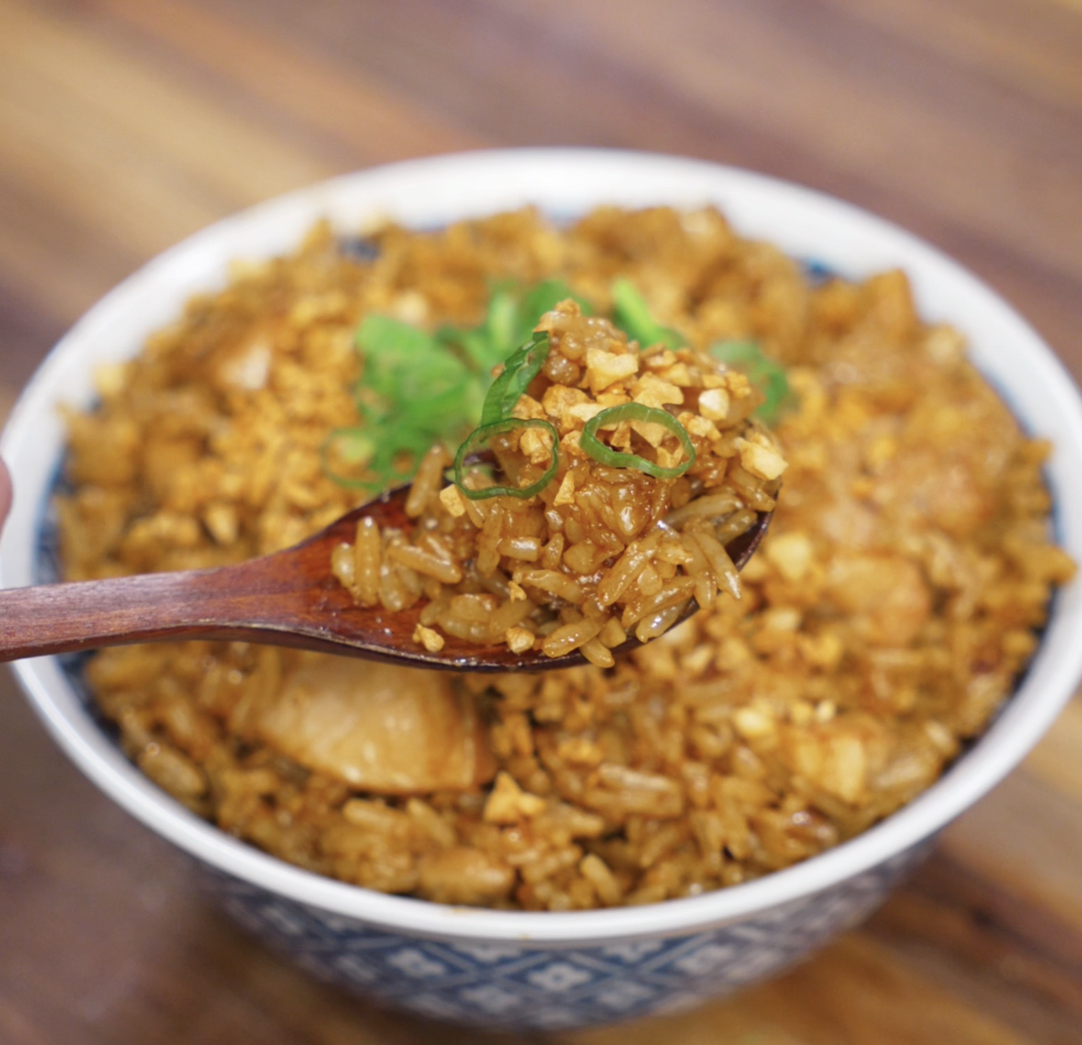 A wooden spoon lifts a bite of garlic chicken fried rice from the bowl, revealing glistening grains of rice, chicken chunks, and crispy garlic in each bite.