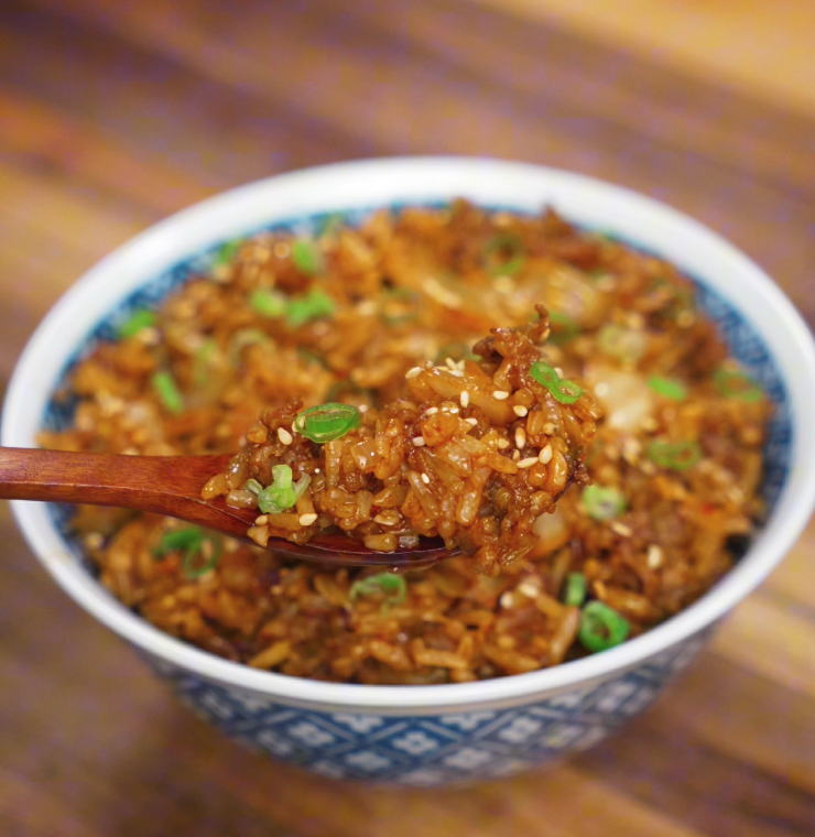 A wooden spoon lifts a generous bite of kimchi beef fried rice, showing glossy grains, caramelized kimchi, and bits of savory beef.