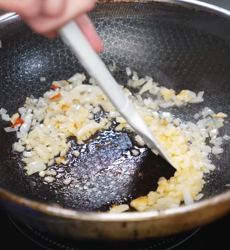 A white spatula stirs diced onion, garlic, and chilies in a hot pan until aromatic and golden, forming the base of the fried rice.