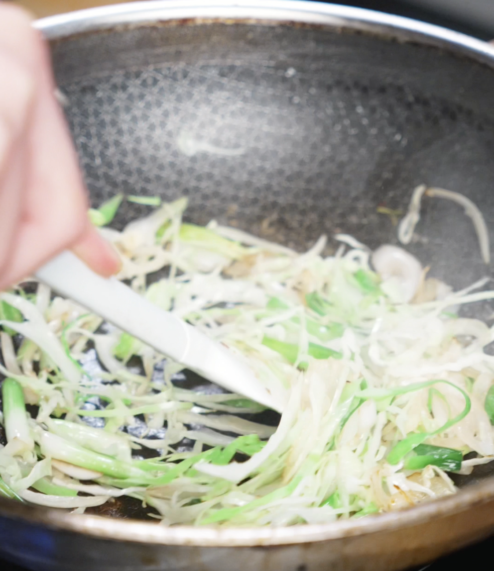 Sliced onions and scallion whites are being sautéed in a wok. These vegetables create an aromatic base for the sauce to cling to later.