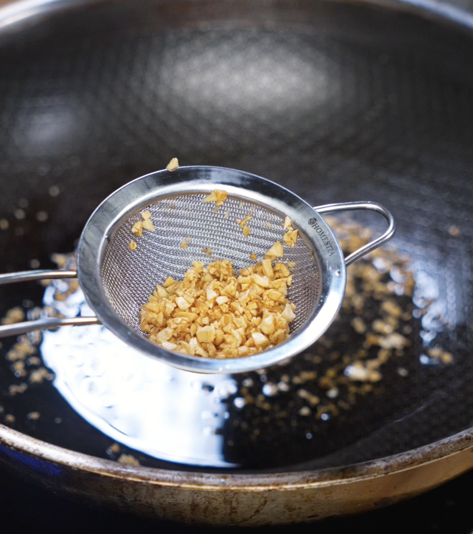 Fried garlic is lifted from a pan of hot oil using a fine mesh strainer, golden and crisp, ready to be used as a topping.