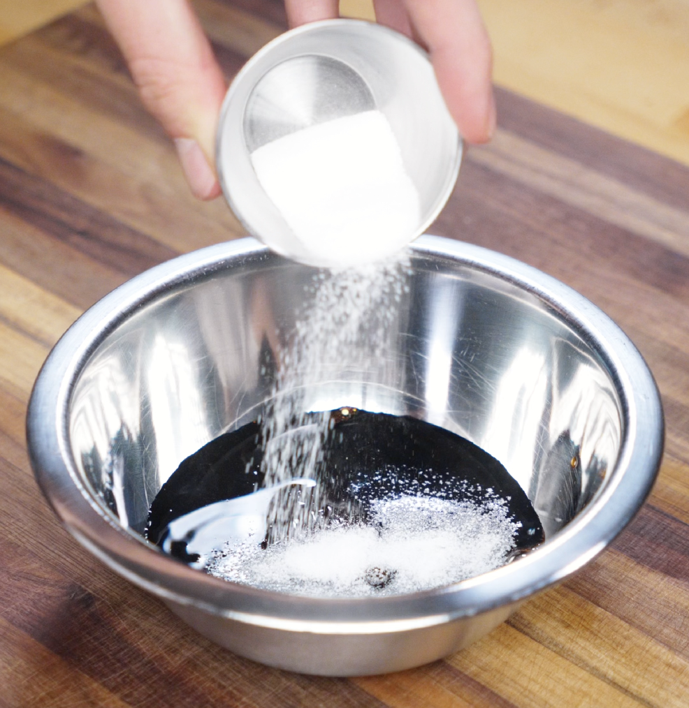 Granulated sugar is being poured into a metal bowl filled with soy sauces and oyster sauce. This simple sauce blend brings deep umami and subtle sweetness to the noodles.