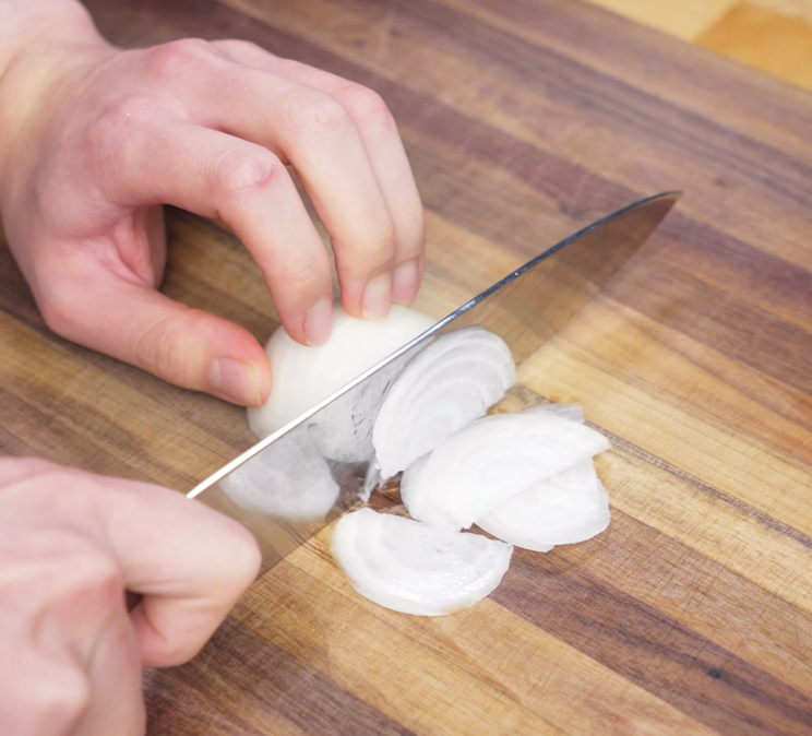 A hand slices a white onion into thin strips on a wooden cutting board, preparing the aromatics for the ginger pork bowl.