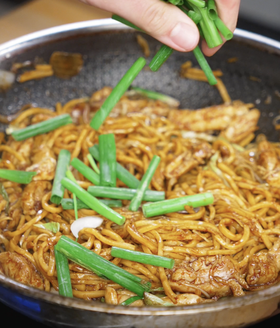 Fresh scallion greens are being added to the pan as the final touch. They add a pop of color, crunch, and brightness to the finished noodles.