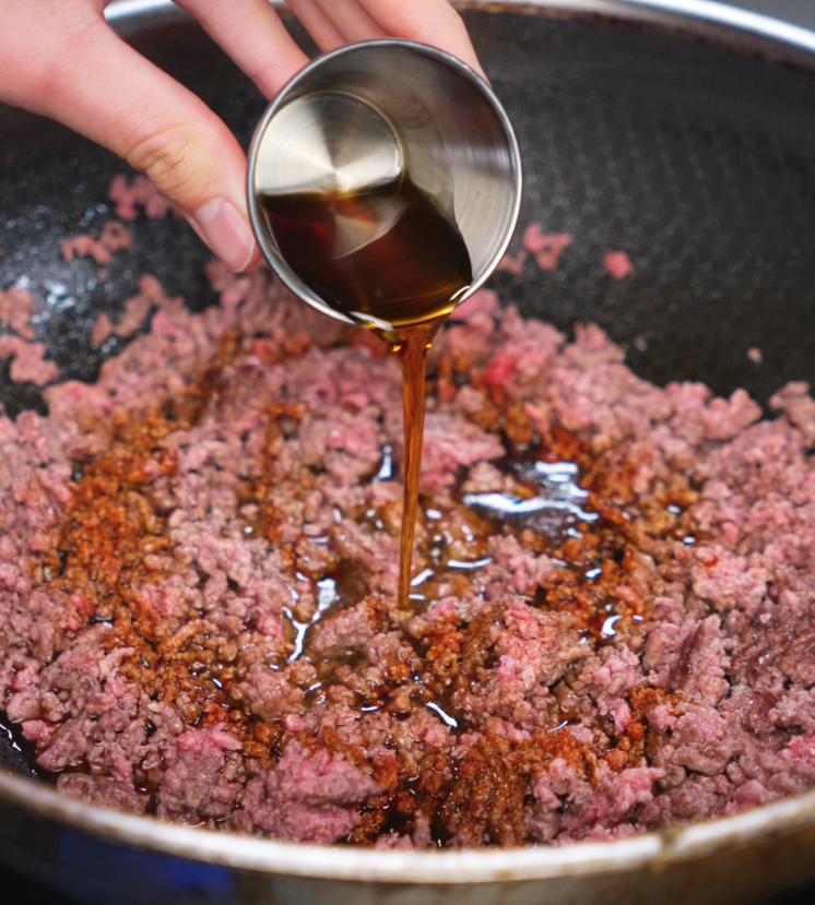 Pouring honey garlic sauce into a pan of browned ground beef. The sweet and savory glaze begins to sizzle and coat the meat as it cooks down.