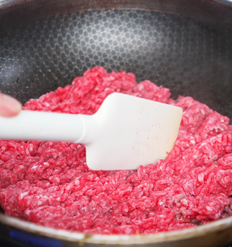 Breaking up raw ground beef in a nonstick skillet with a white spatula to begin cooking honey garlic beef. The beef is starting to brown and cook evenly.