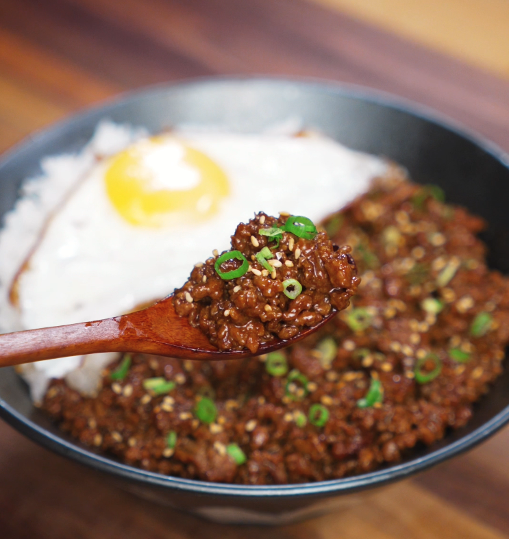 Wooden spoon lifting a flavorful bite of honey garlic beef over the rice bowl. The saucy beef is rich and glossy, with egg and rice in the background.