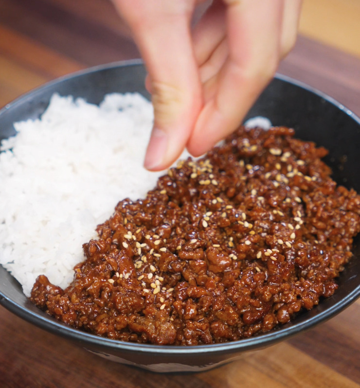 Garnishing a honey garlic beef rice bowl with toasted sesame seeds. The glossy beef is served next to a bed of white rice in a black bowl.