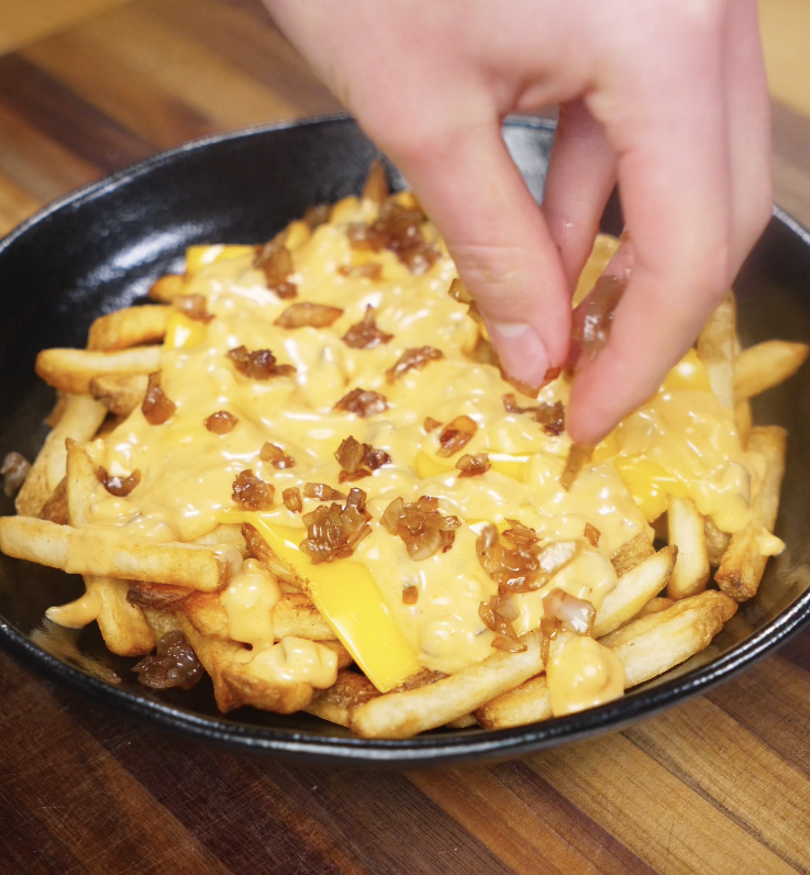 A hand sprinkles sweet caramelized onions over the cheesy fries for extra flavor and texture.