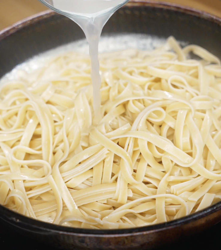 A splash of reserved pasta water is poured into the pan of cooked fettuccini to loosen and emulsify the Alfredo sauce.