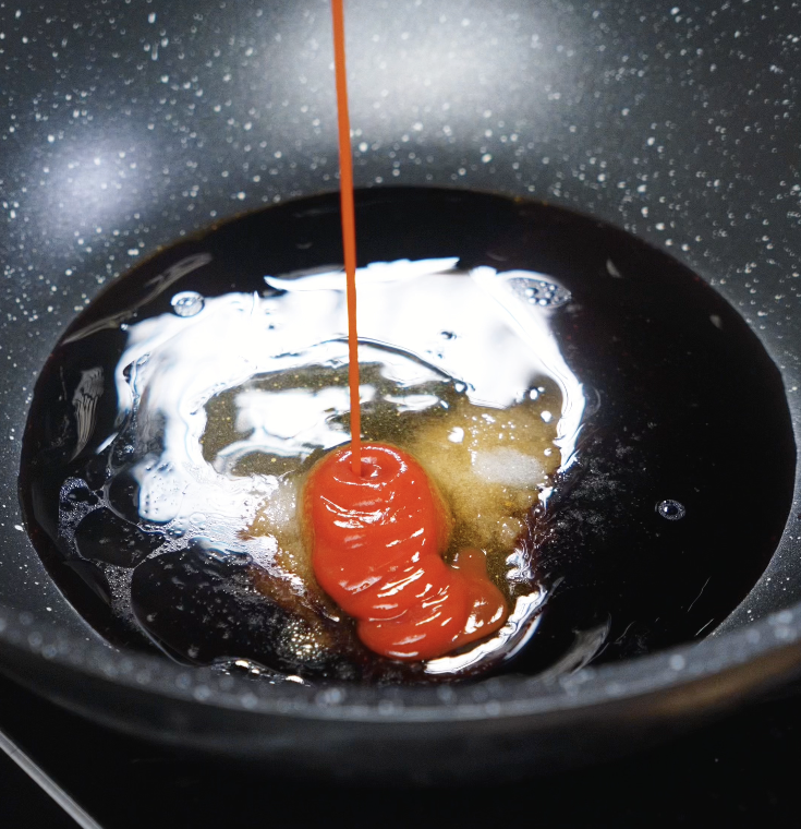 Thick ketchup being poured into a mixture of soy sauce, vinegar, sugar, and sesame oil in a nonstick pan.