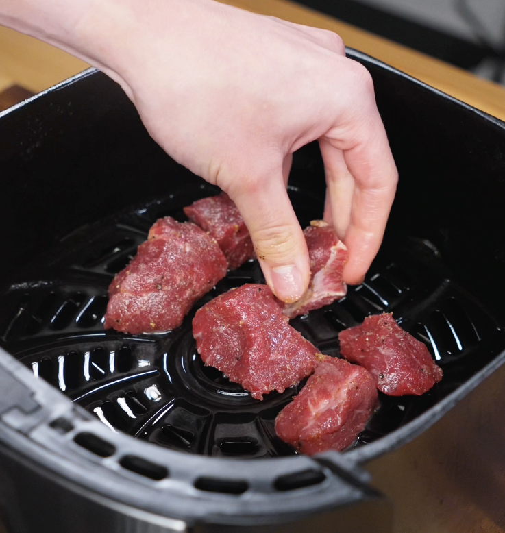 Raw steak cubes are being arranged in an air fryer basket, spaced evenly before cooking.