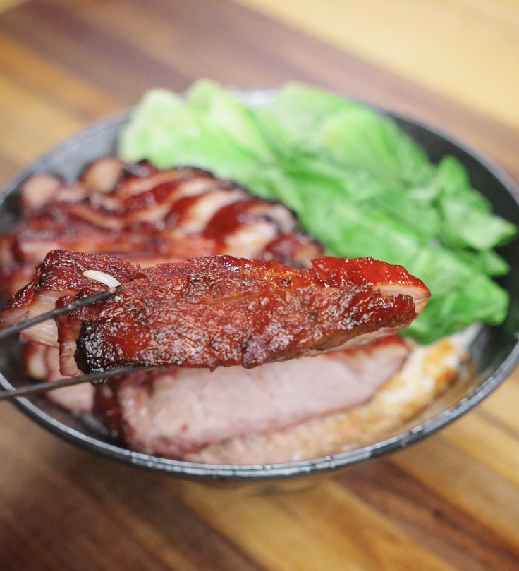 A juicy slice of char siu pork held up with chopsticks, showing its tender interior and caramelized edges.