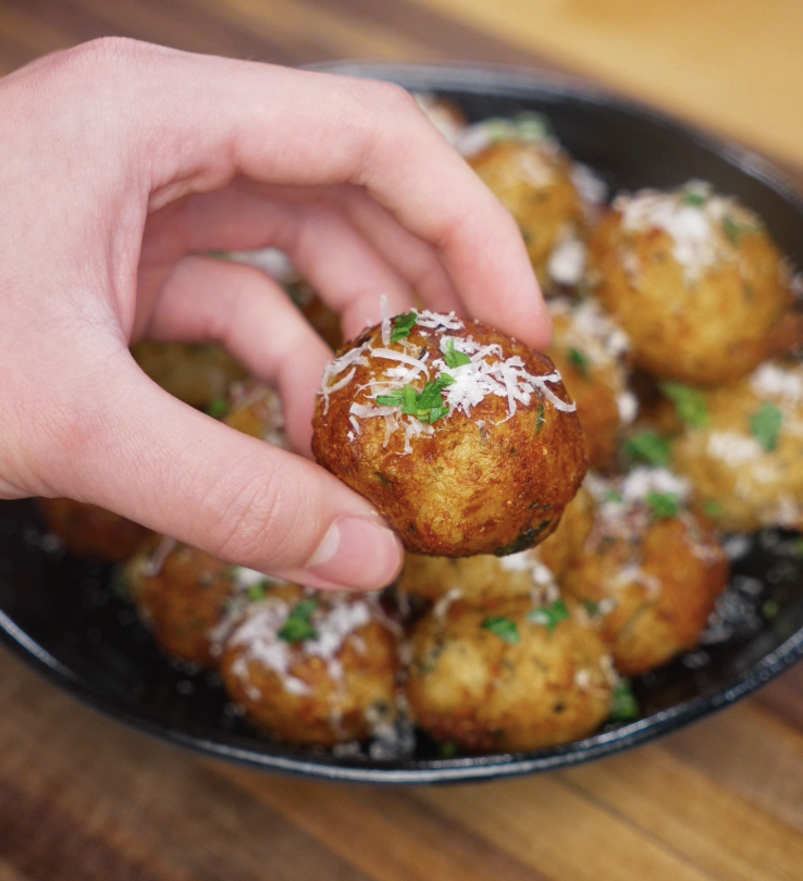 A single crispy potato ball is held up by hand, showcasing its golden brown crust and sprinkling of parmesan and herbs.